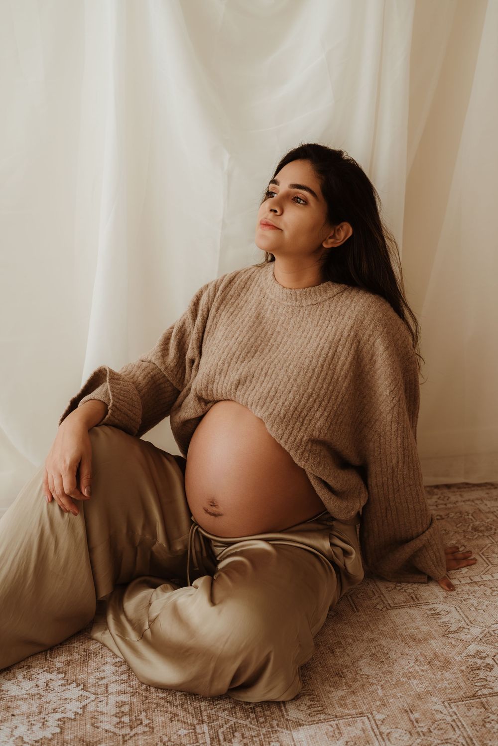 Artistic maternity photo series showing pregnant person in beige sweater reclining against white backdrop.