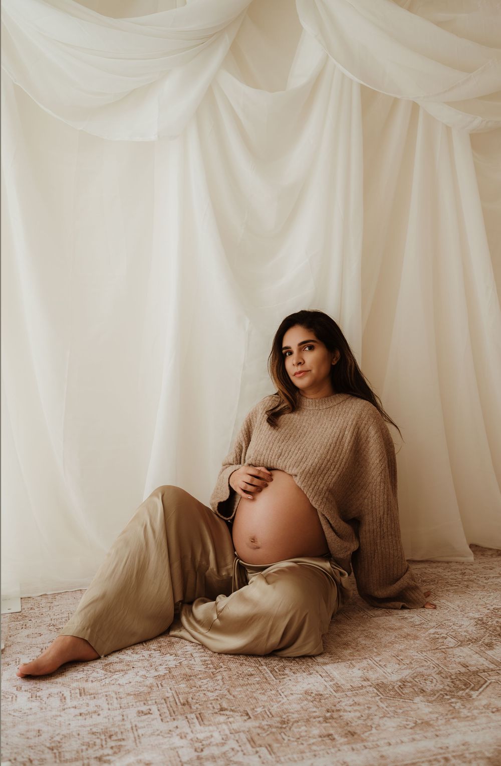 A series of maternity photos showing a pregnant woman in a beige sweater posing against a white draped backdrop.