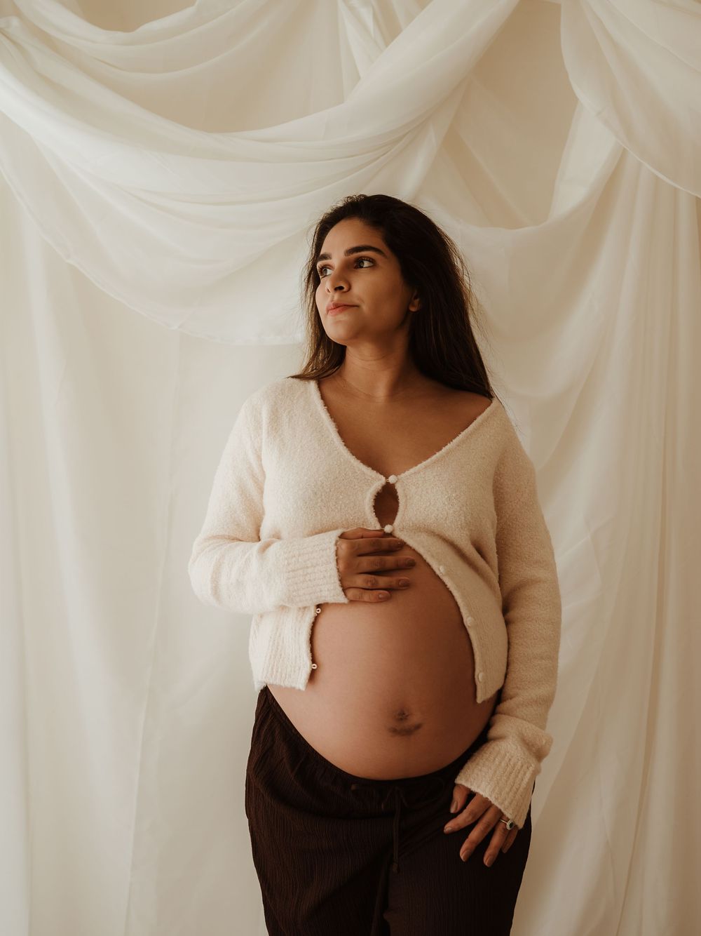Expectant mother in cream sweater poses against white draped fabric backdrop during maternity photoshoot.