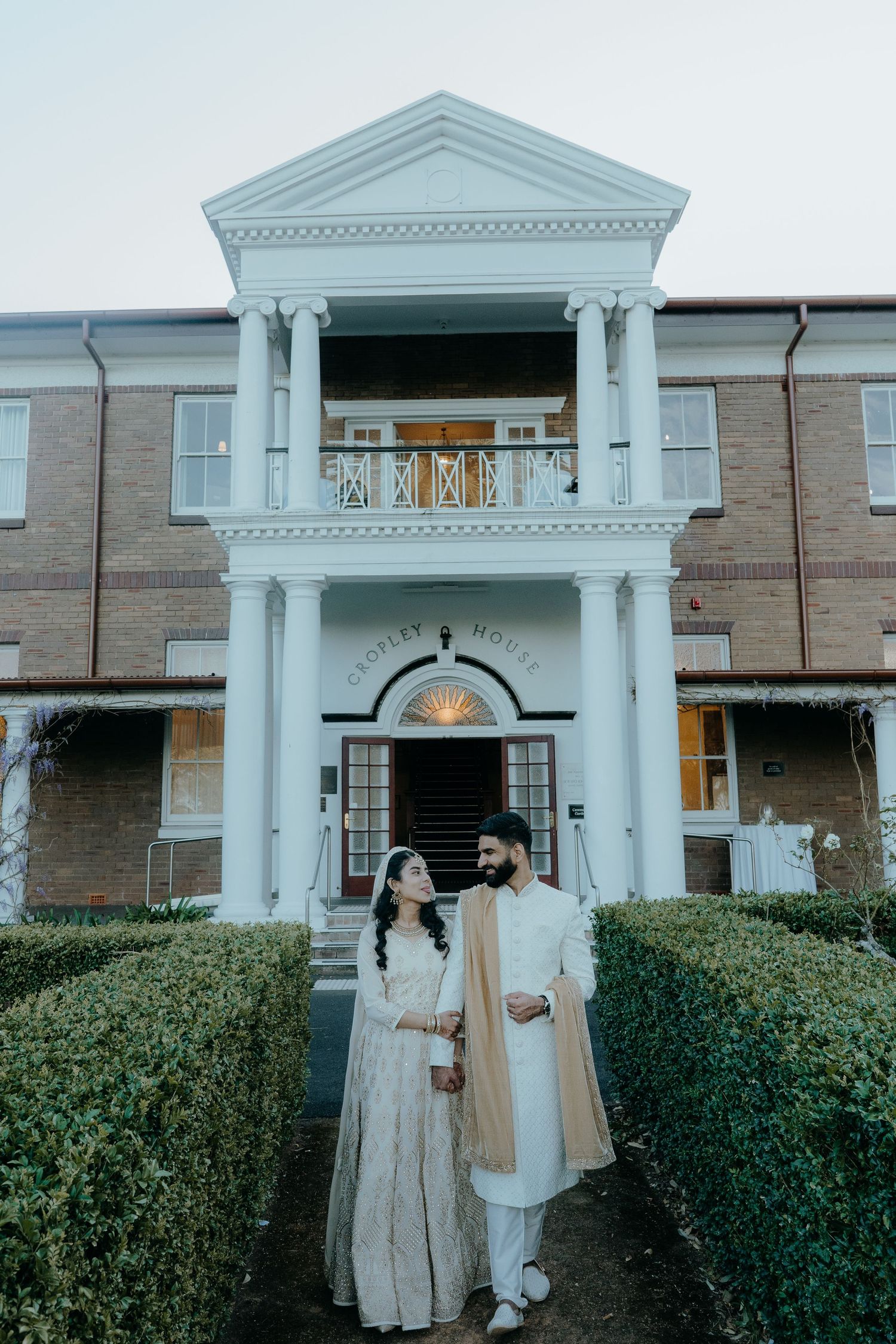 Traditional South Asian couple in wedding attire poses in front of a stately white columned building entrance.