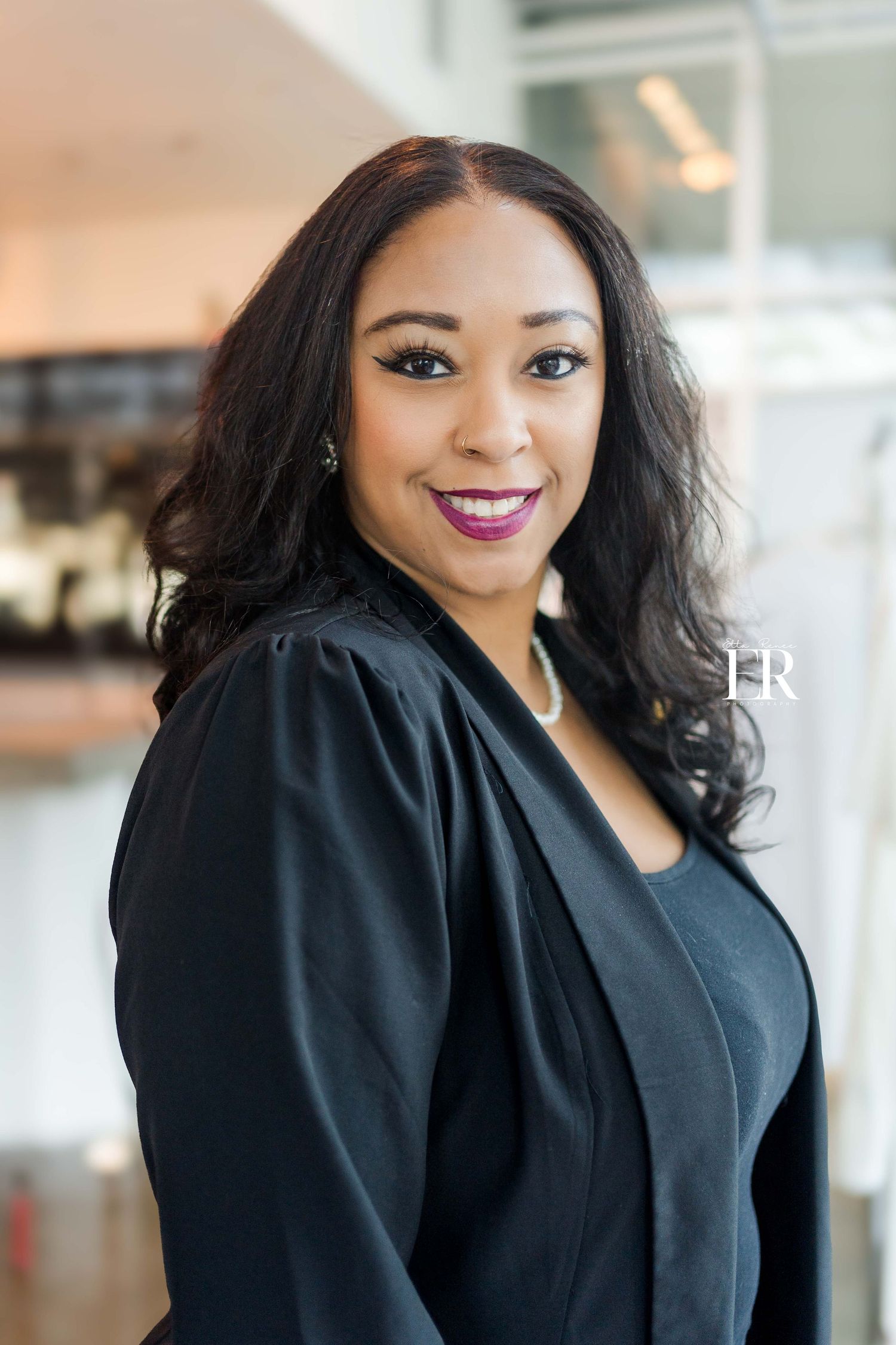 Professional portrait of woman in black attire with styled hair and makeup in office environment.