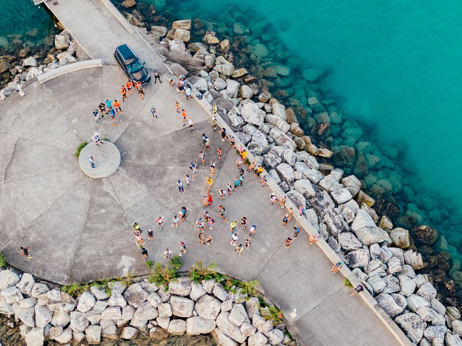 Aerial view of people gathering on a concrete pier with turquoise waters and large rocks along the coastline.