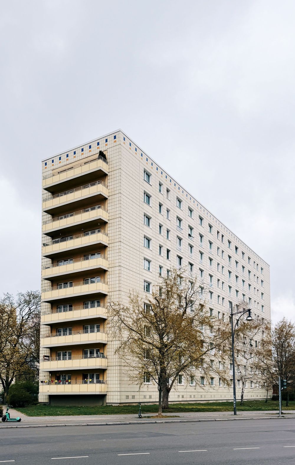 A tall modernist apartment building with beige facade and distinctive stacked balconies stands against a cloudy sky.