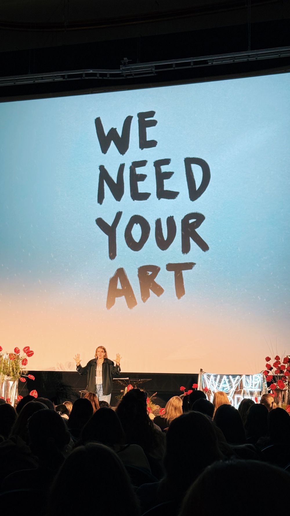 Text saying 'WE NEED YOUR ART' displayed on stage screen with crowd silhouettes at an event.