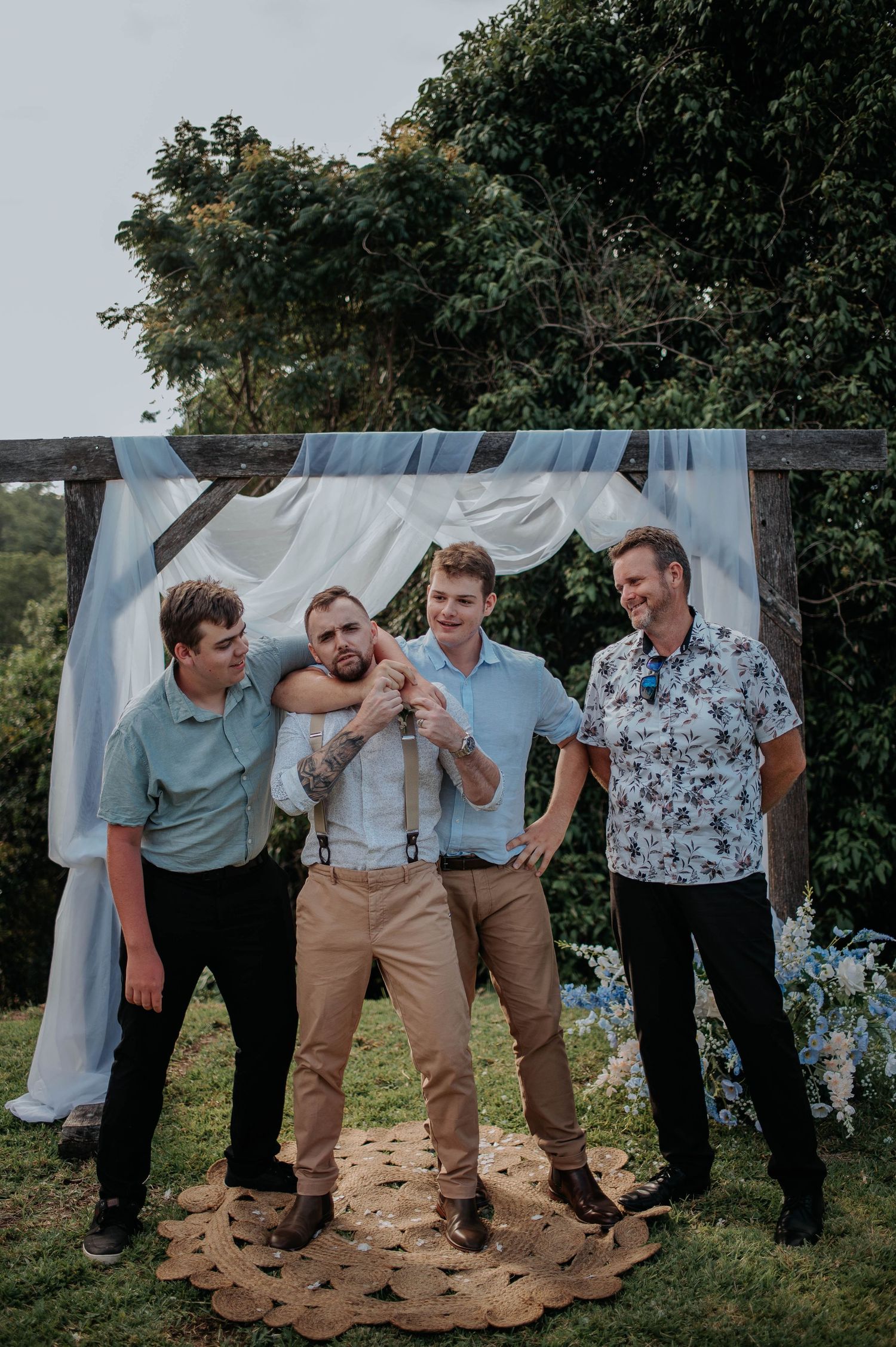 A group of men in casual attire laughing together at an outdoor wedding ceremony with white drapes in the background.