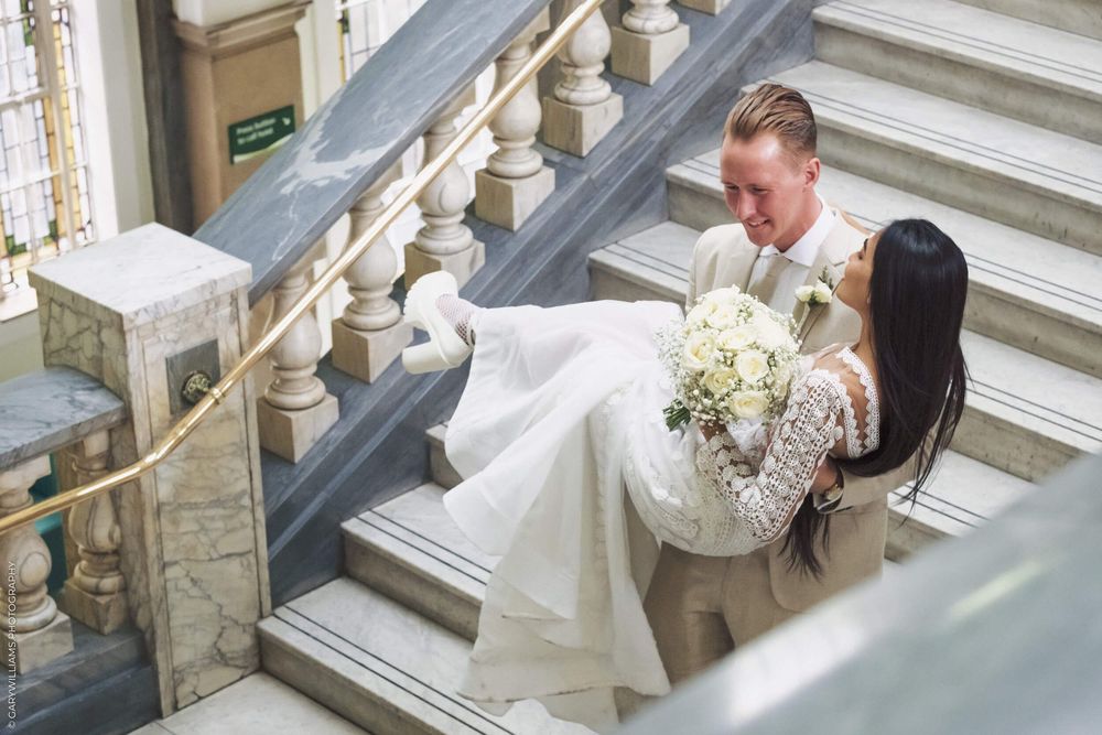 Two people in wedding attire share a romantic moment on elegant stone stairs with white bridal bouquet in hand.