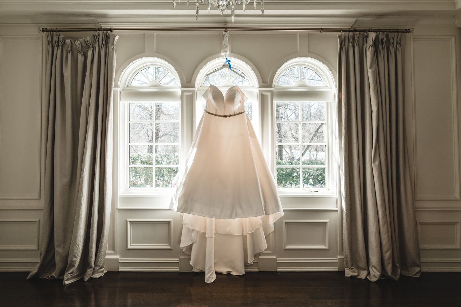 Wedding dress hangs in front of elegant windows with natural light streaming through.