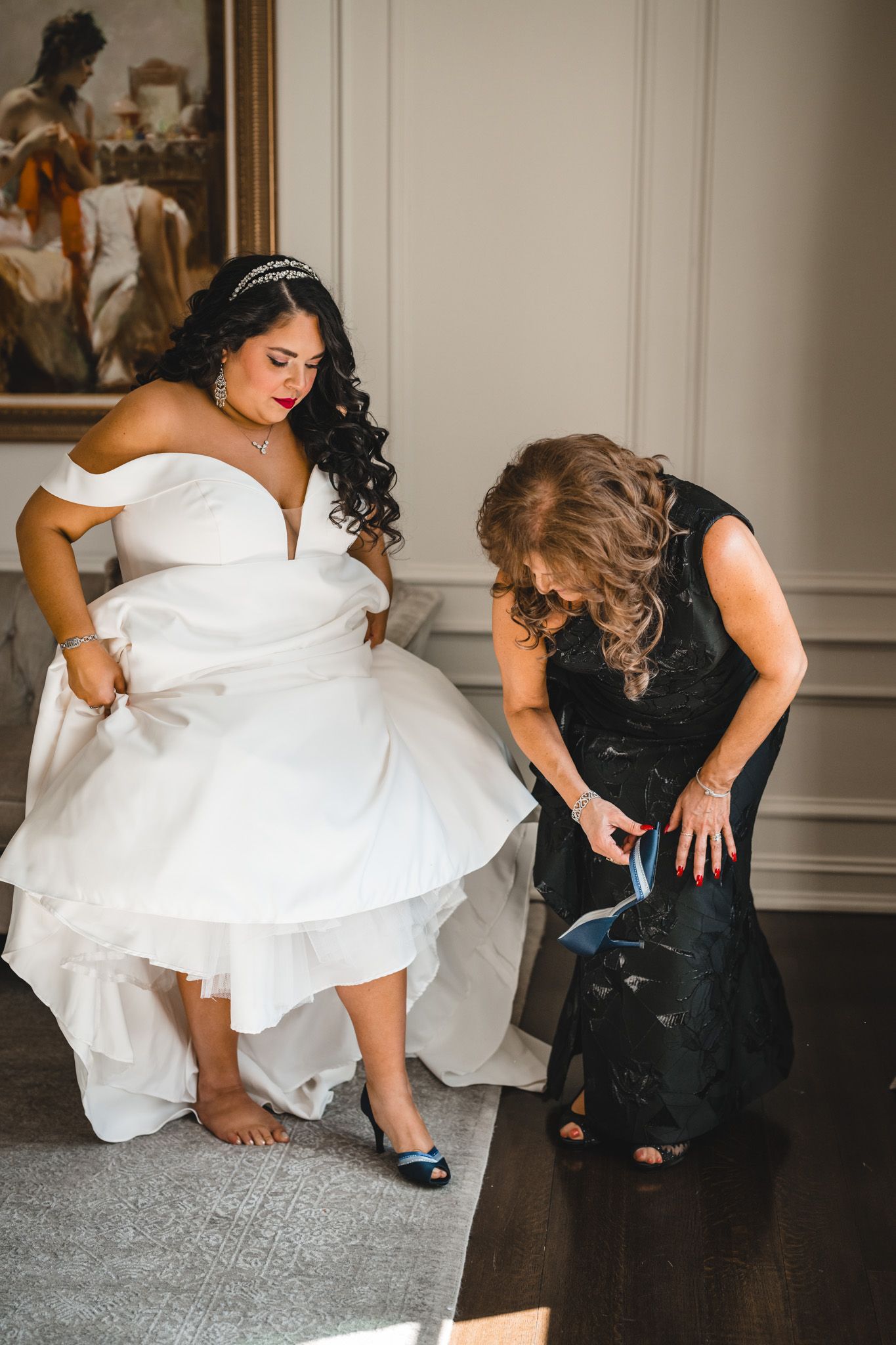 Wedding preparations showing a bride in white dress getting help with her shoes from someone in black.