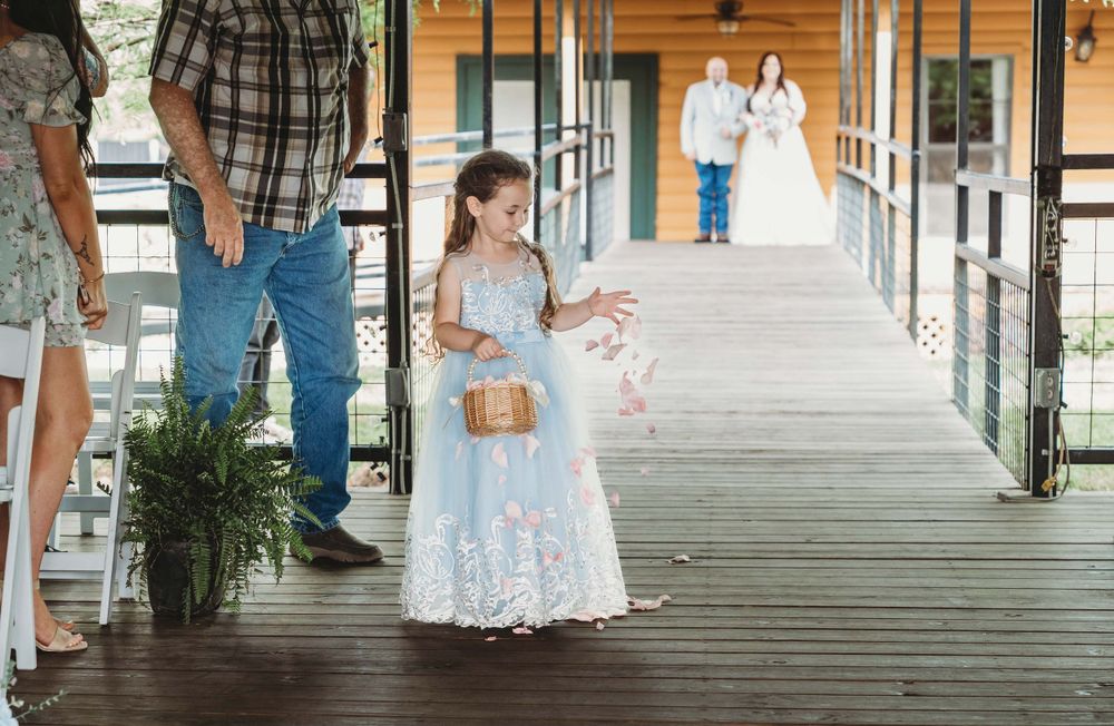 A flower girl in a pale blue lace dress walks on a covered wooden porch with her flower basket.
