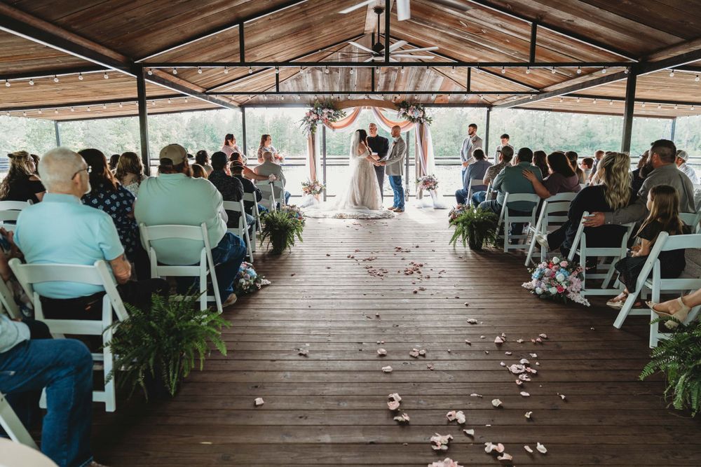 Wedding ceremony taking place on a wooden deck pavilion with white chairs and scattered rose petals down the aisle.