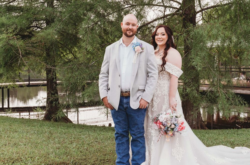 A couple poses outdoors at their wedding ceremony near pine trees and a lake in a rustic outdoor setting.