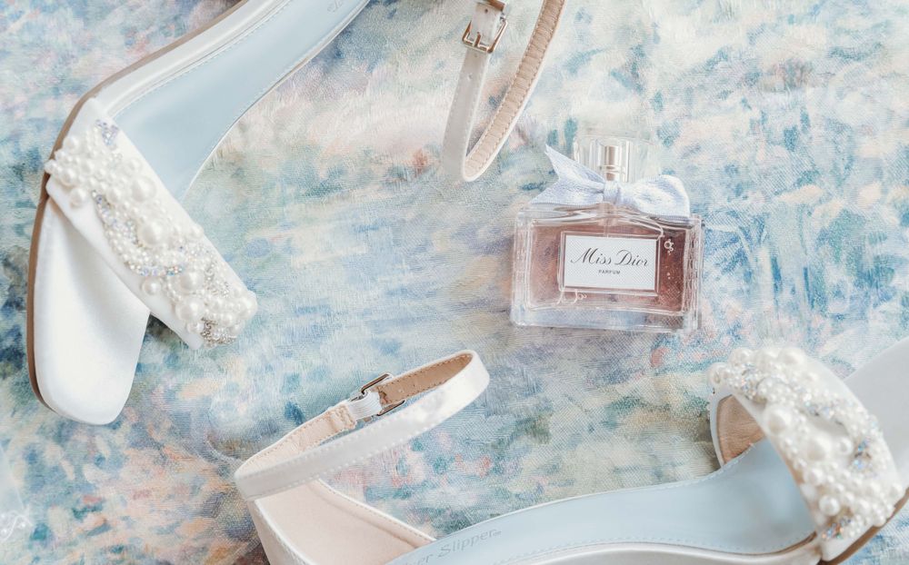 Elegant bridal accessories including white high heels, lace garter, and pink perfume bottle arranged on light blue surface.