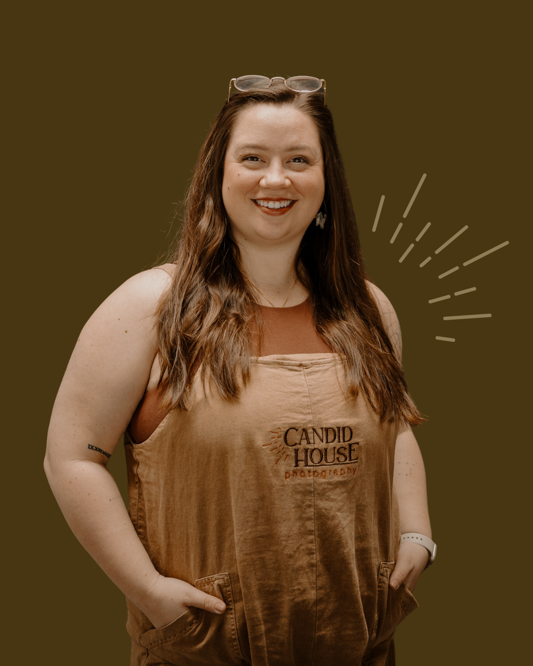 A smiling person wearing a brown Candid House tank top and sunglasses on their head stands against a dark background.
