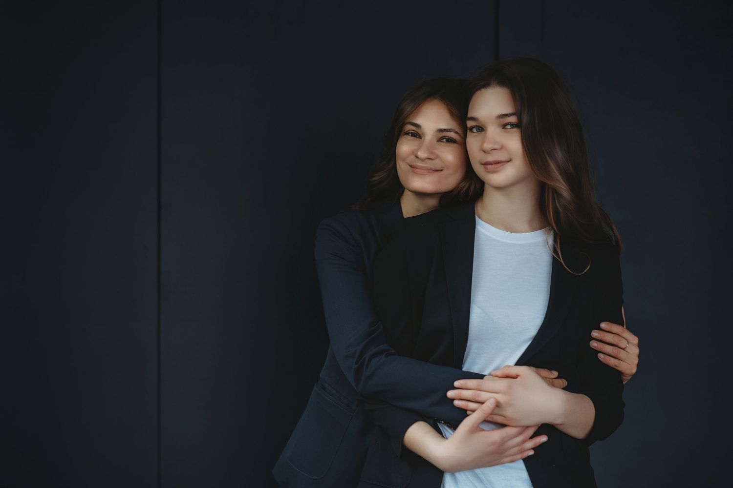 Two people share a warm embrace and smile while standing against a dark wall.