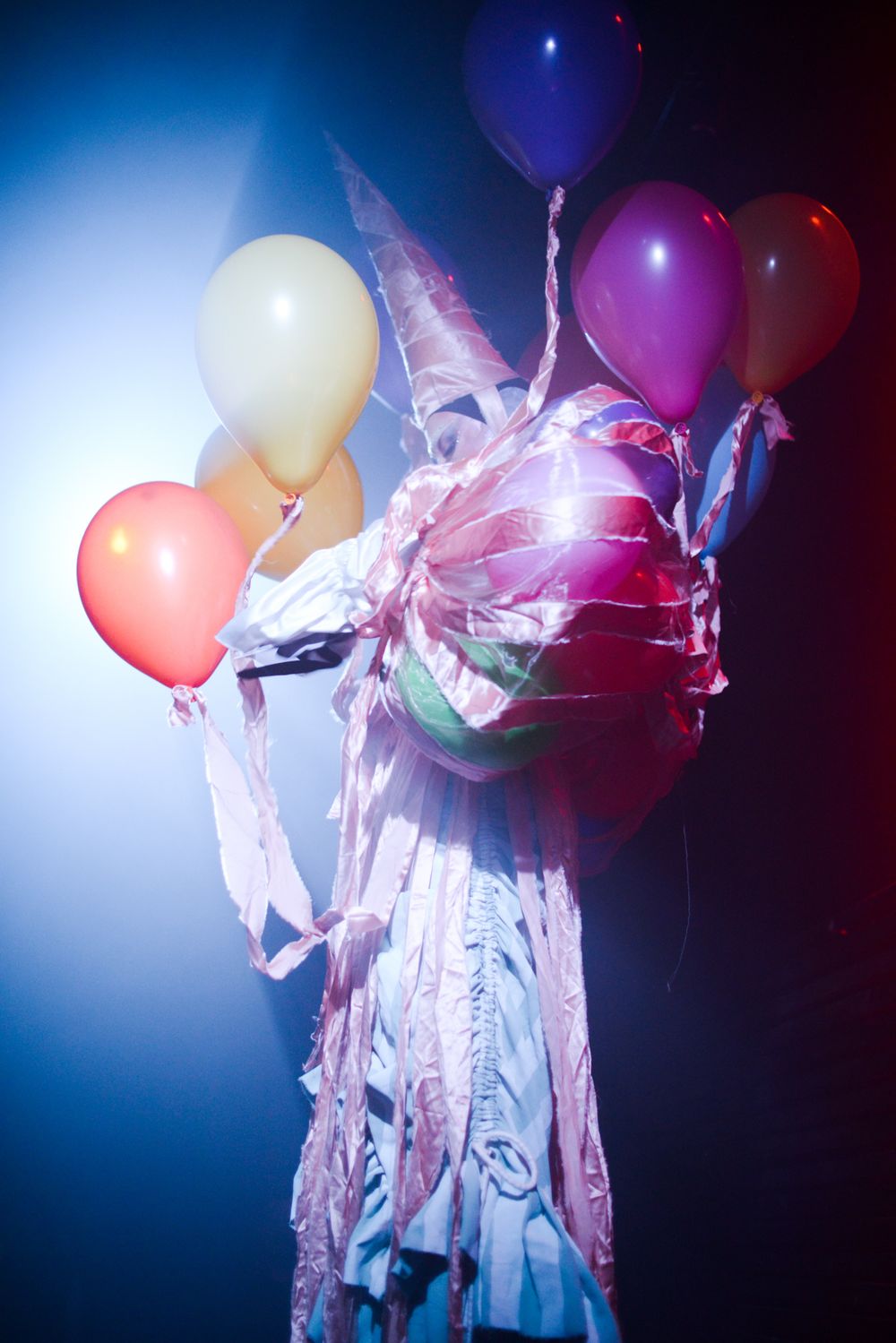 Artistic shot of a figure in white fabric holding colorful balloons against blue lighting.