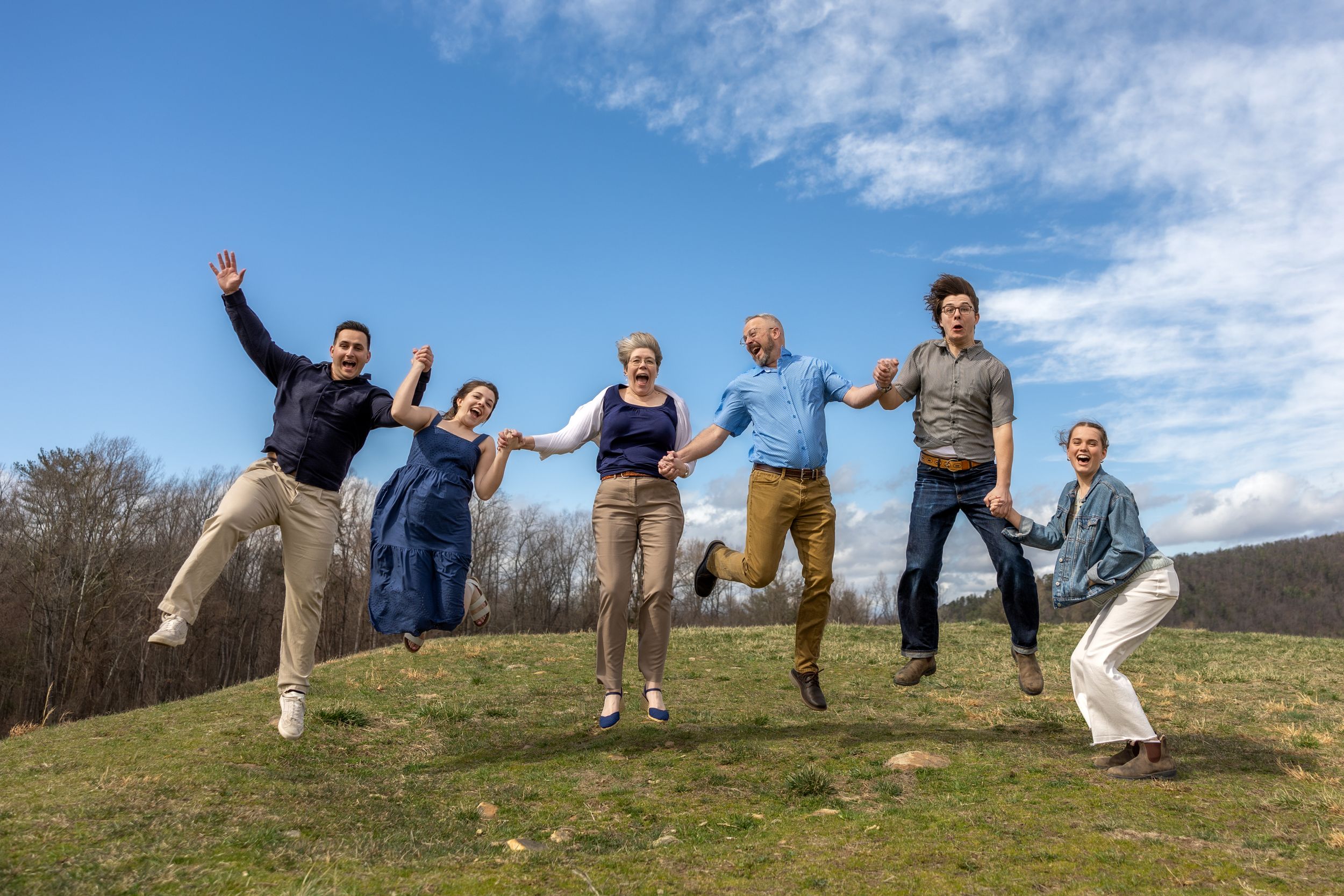 A group of people jumping joyfully on a grassy hilltop against a blue sky with white clouds.