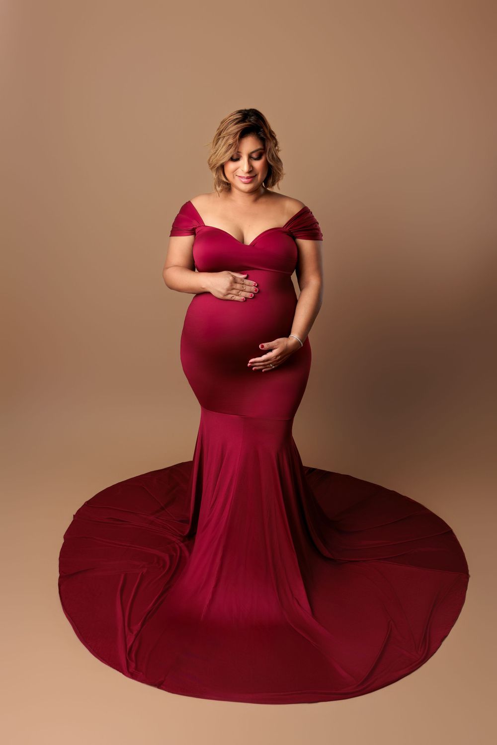 Stunning maternity photo of an expectant mother in a flowing burgundy off-shoulder gown against neutral background.
