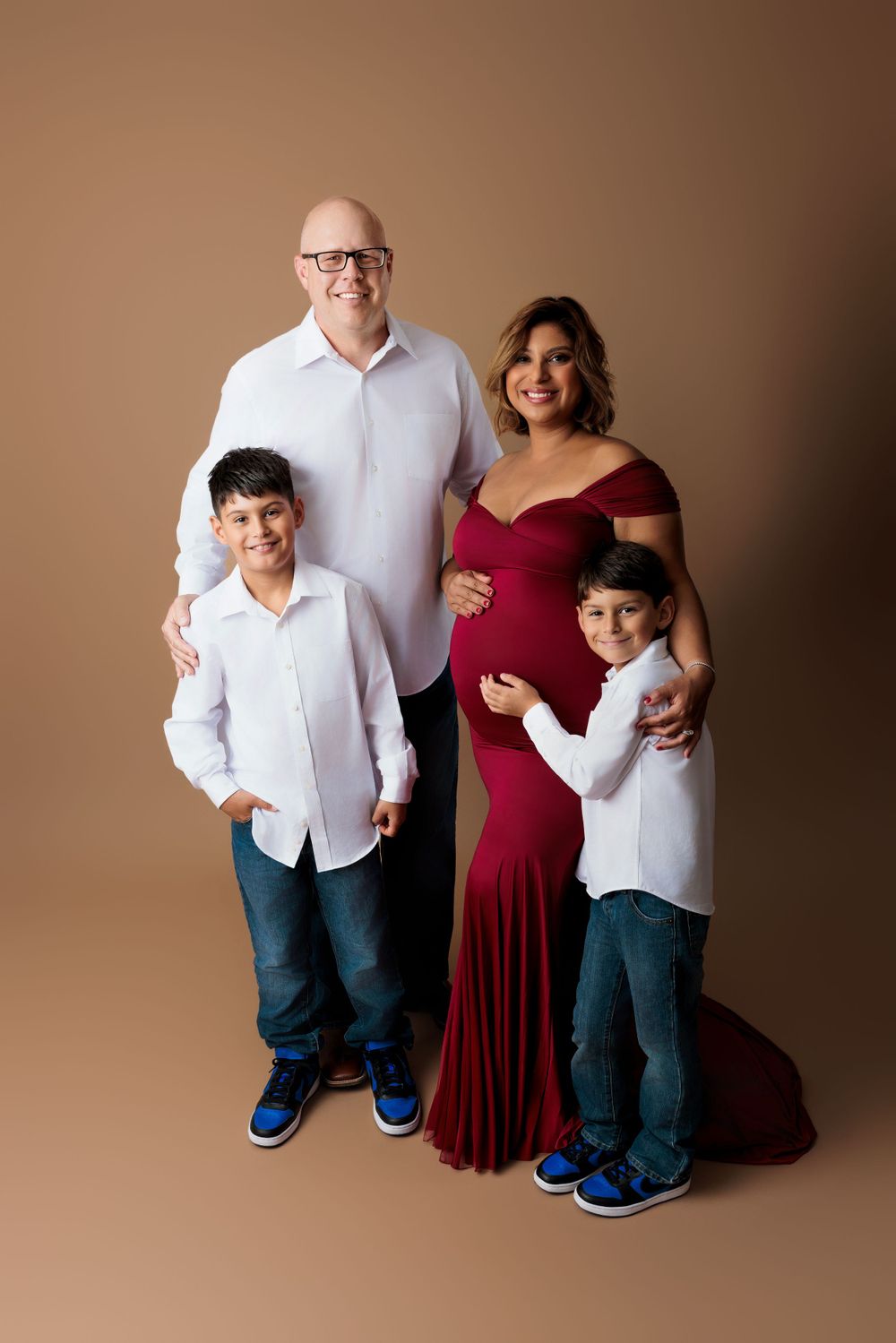 A family of four poses for a studio portrait against a brown backdrop, wearing coordinated white and red attire.