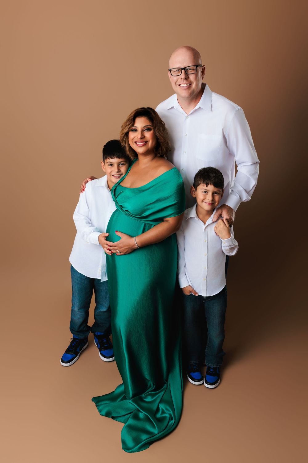 A family of four poses in coordinated outfits against a brown backdrop, with the mother wearing an elegant emerald green gown.