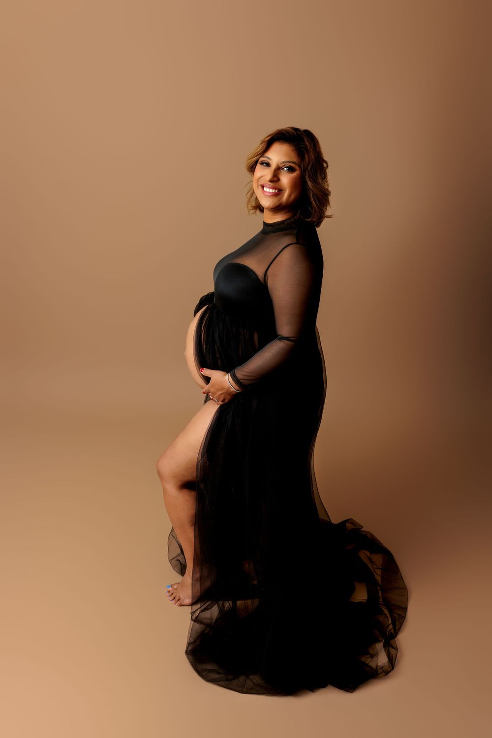 Maternity photoshoot featuring an elegant black sheer gown with dramatic train against a warm beige backdrop.