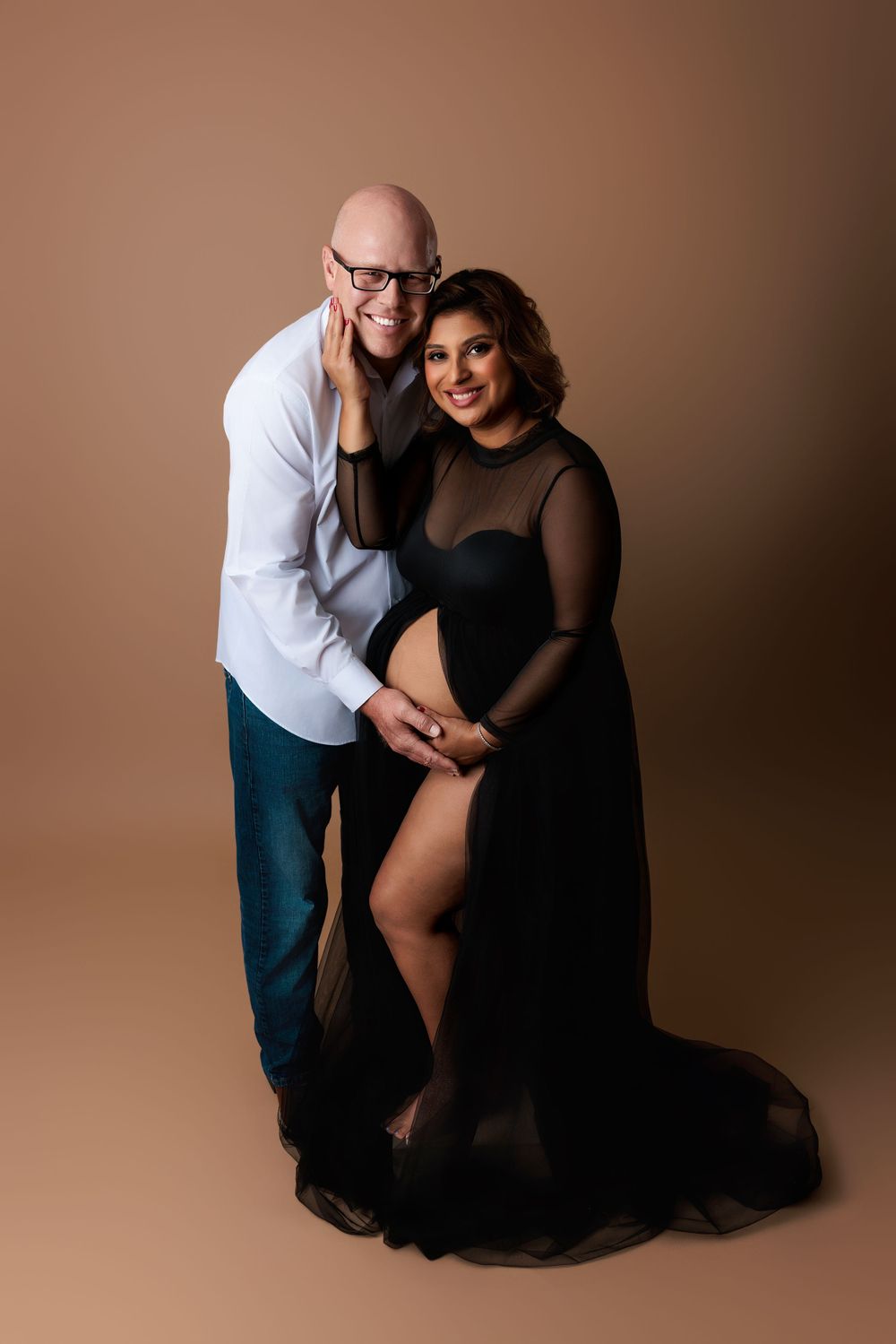 Studio portrait of a couple posing together against brown backdrop, woman wearing elegant black sheer gown.
