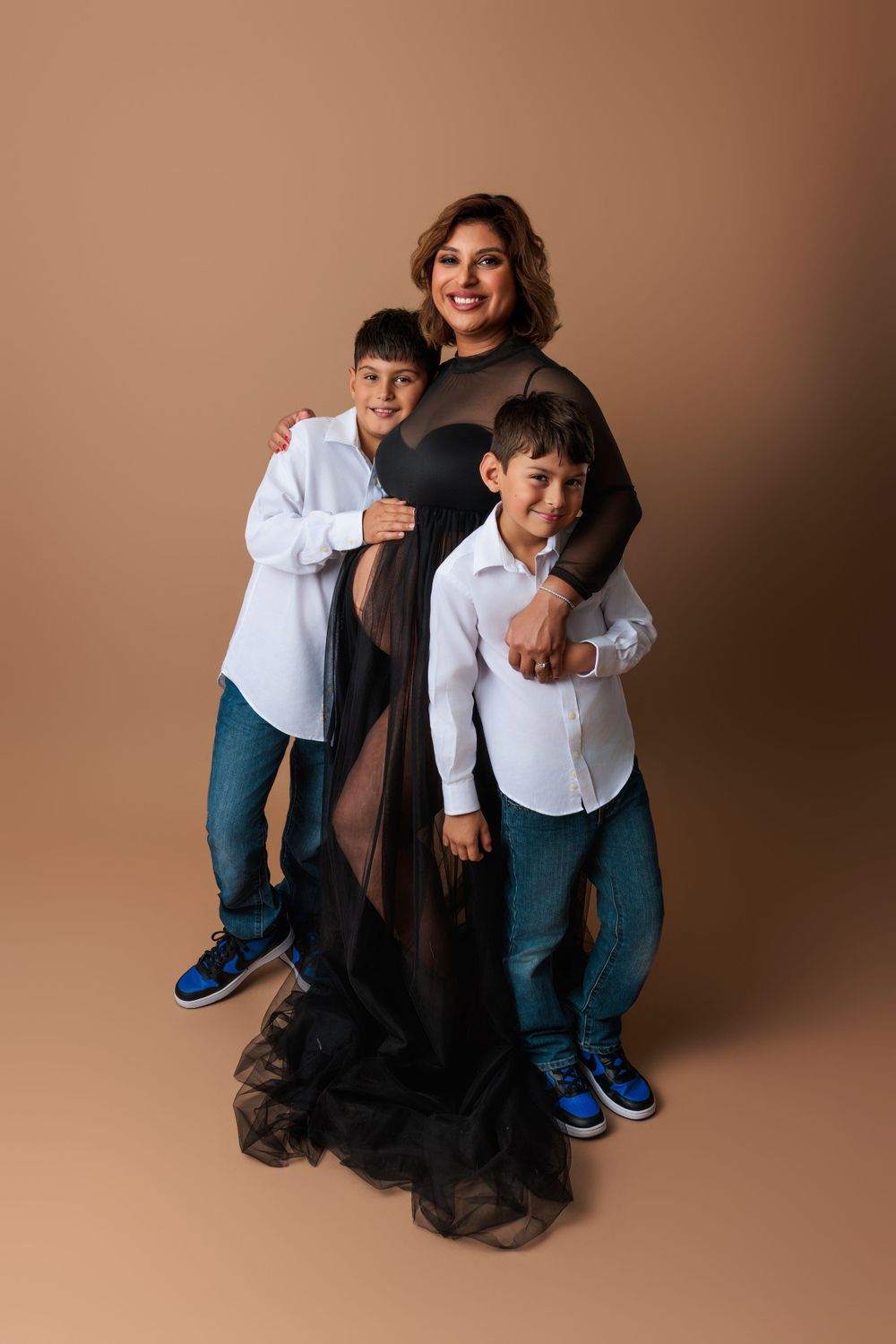 Family portrait against brown backdrop with two children in white shirts and blue sneakers with parent in flowing black dress.