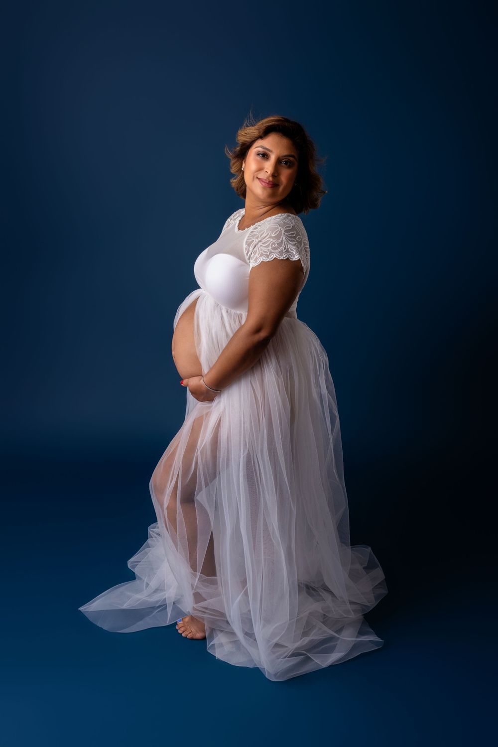 Elegant maternity portrait in flowing white lace gown against deep blue studio backdrop.
