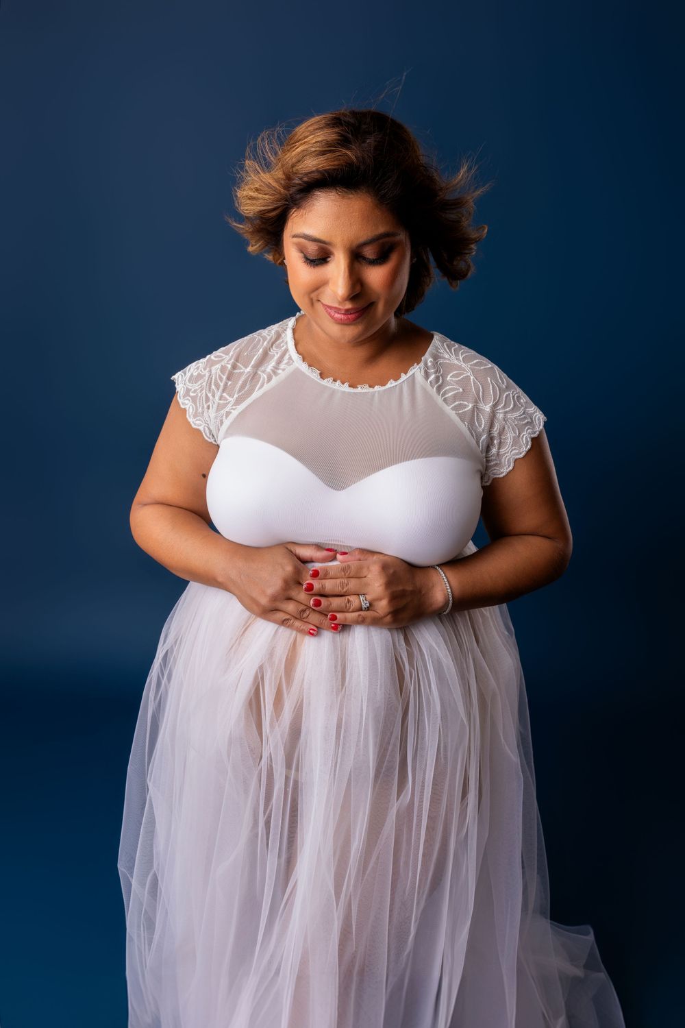 Plus size bride in white lace and tulle dress poses elegantly against deep blue backdrop during bridal photoshoot.