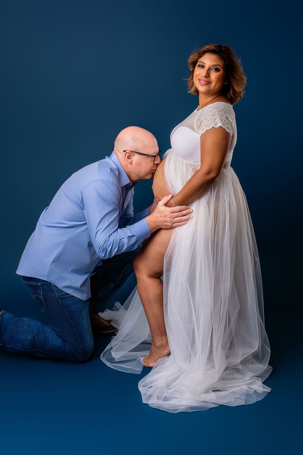 A dreamy maternity photoshoot featuring a couple in soft lighting against a deep blue backdrop.