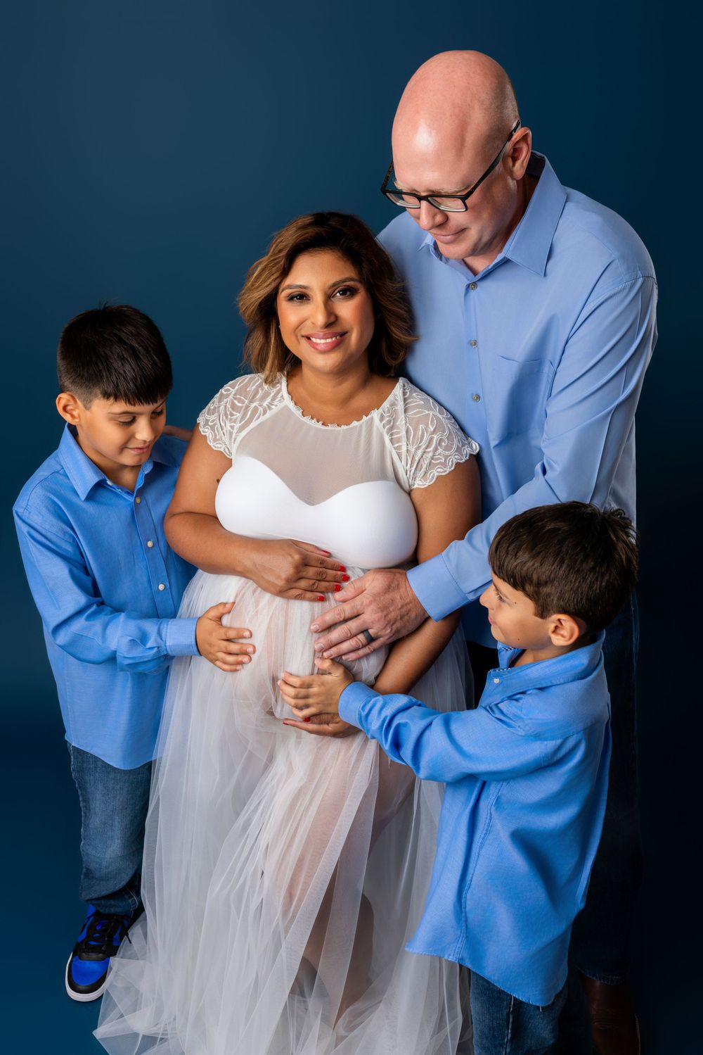 Family maternity portrait with expectant mother in white dress surrounded by loved ones against blue backdrop.