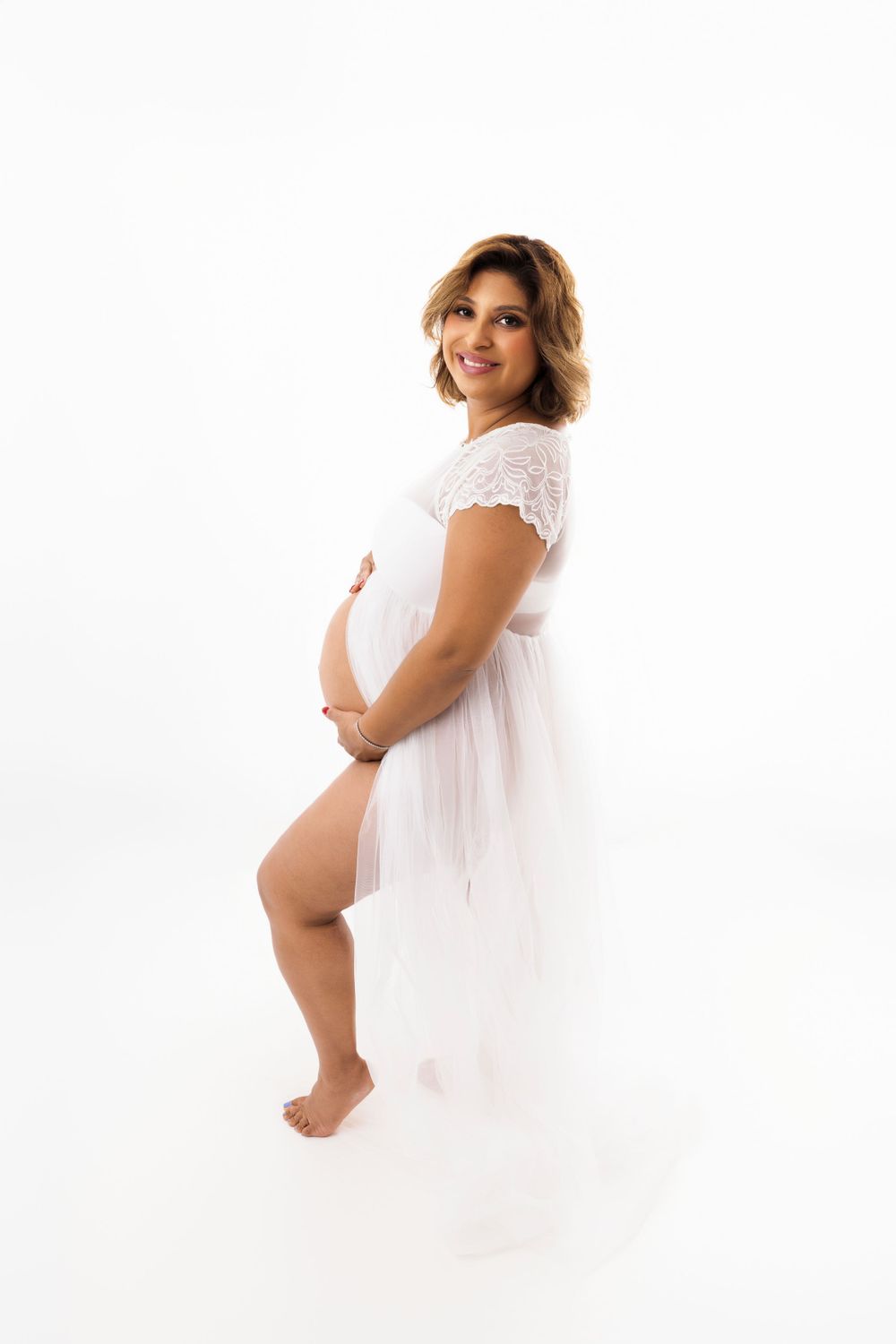 Expectant person in white lace dress poses gracefully on white backdrop during maternity photoshoot.