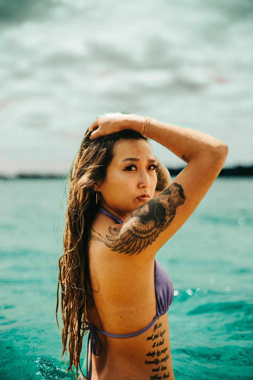 Woman with wing tattoo poses artistically in turquoise ocean waters.