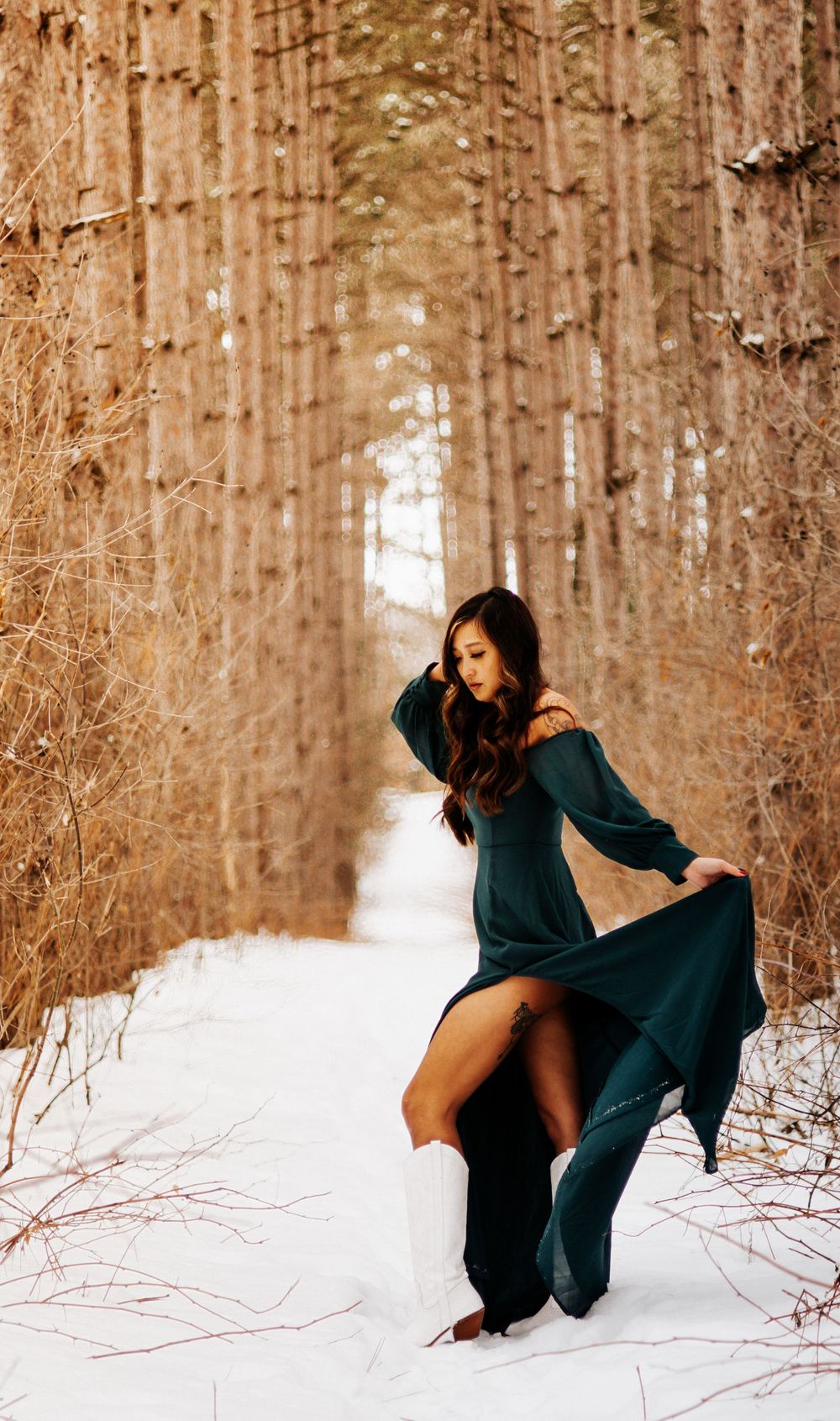 A flowing dark green dress billows dramatically in a snowy forest path lined with tall pine trees.