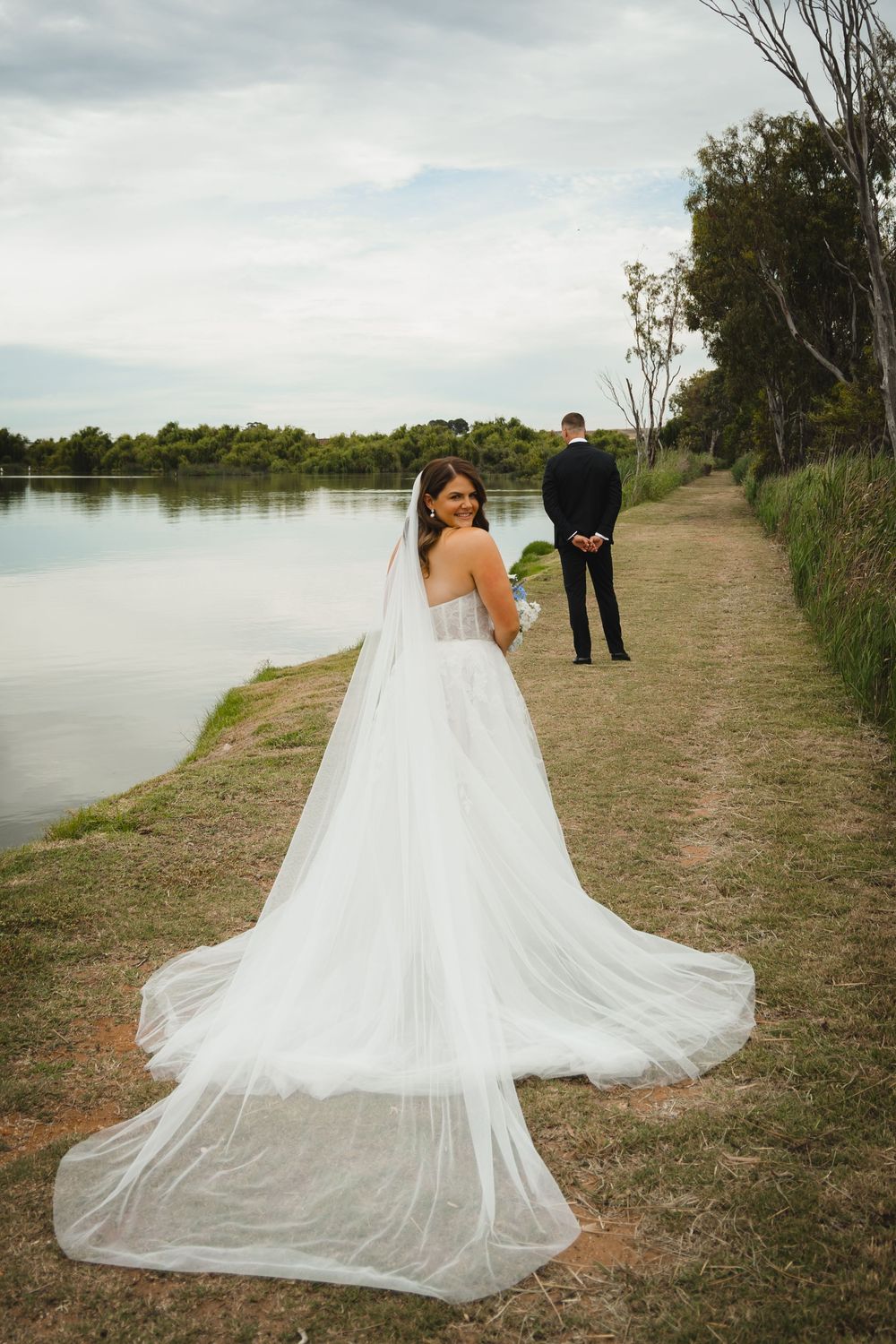 A couple shares a romantic first look moment on a lakeside path with dramatic clouds overhead and flowing white dress.