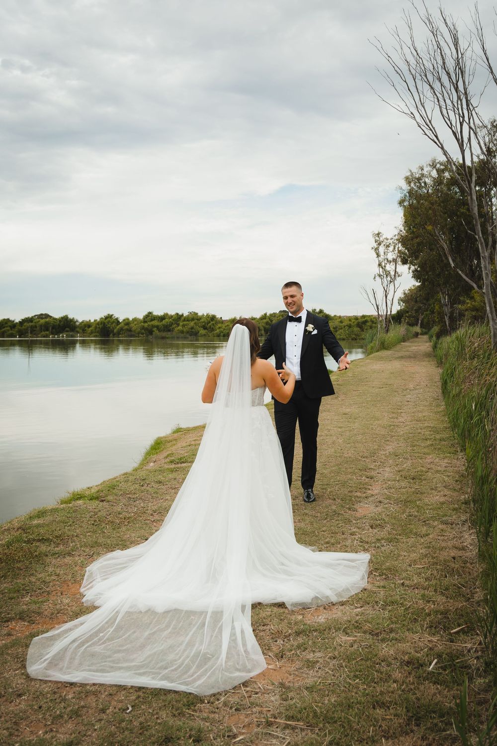 Wedding couple walking along scenic lakeside path with bride's long flowing veil trailing behind them.