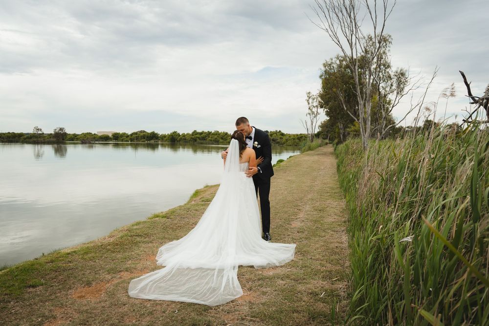 A couple in wedding attire stands along a scenic lakeside path surrounded by tall reeds and calm waters.
