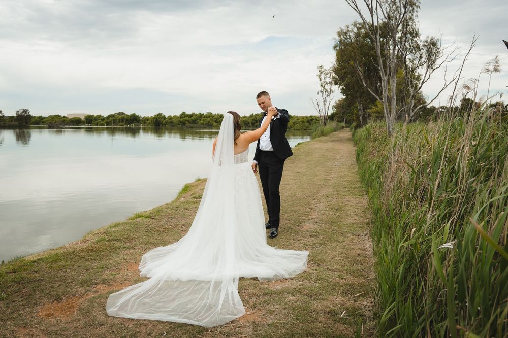 A bride and groom embrace on a dirt path beside a serene lake with tall grasses on a cloudy day.
