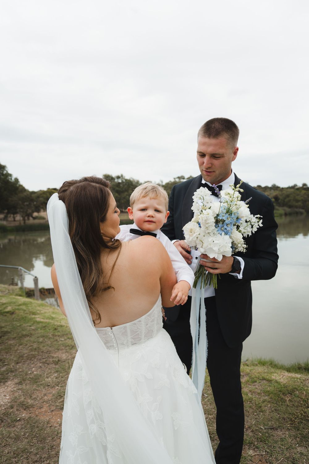 A couple and toddler stand by a lake during a wedding ceremony with white floral bouquet.