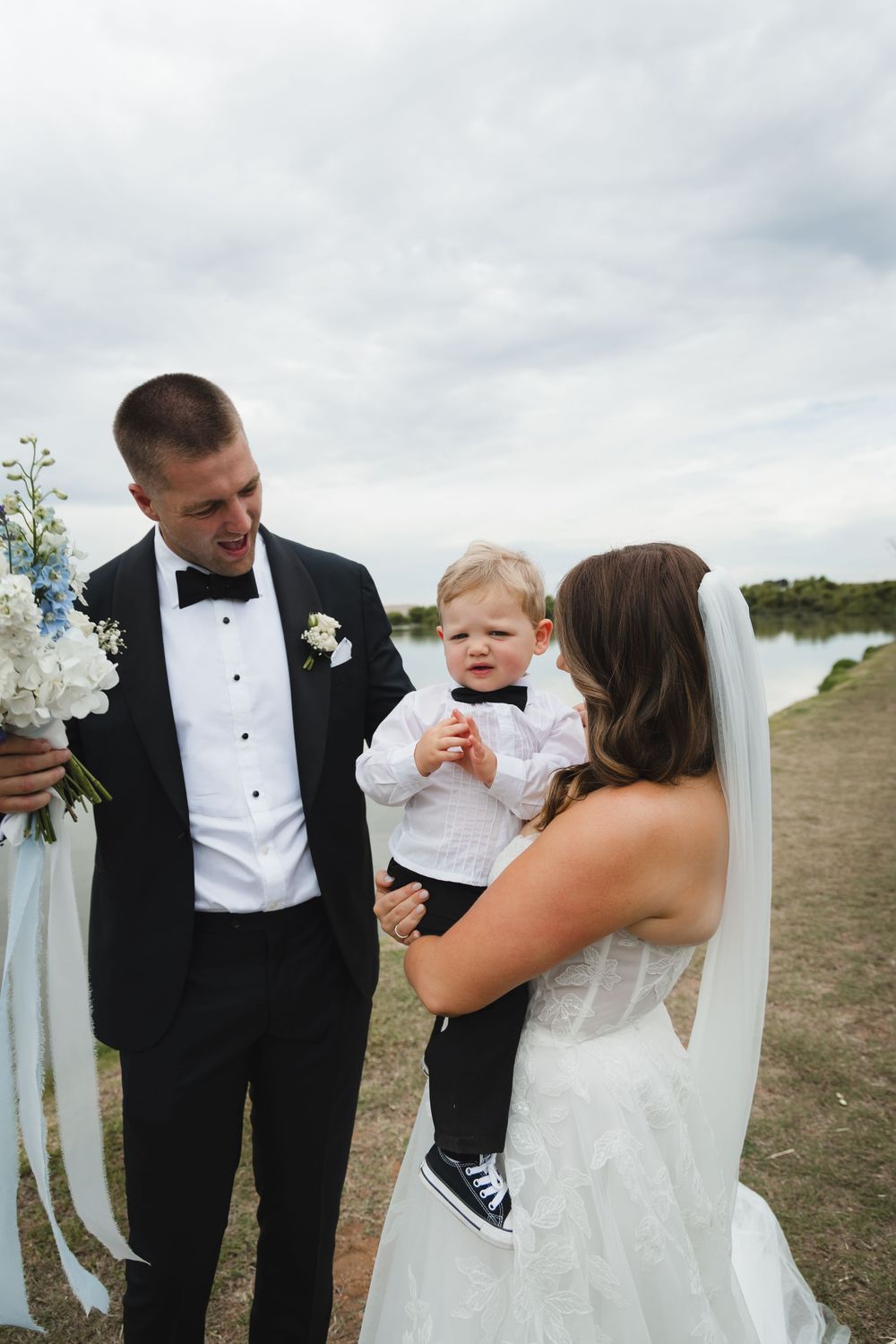 A wedding couple and a small child in formal attire share a joyful moment outdoors on their special day.