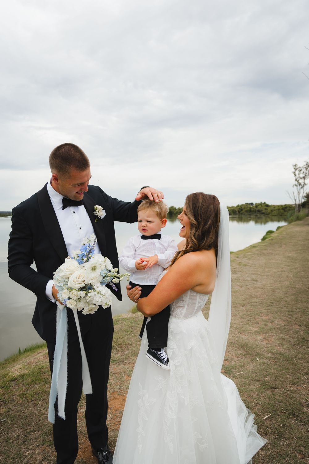 Elegant wedding couple shares a sweet moment with a child by a lakeside on their special day.