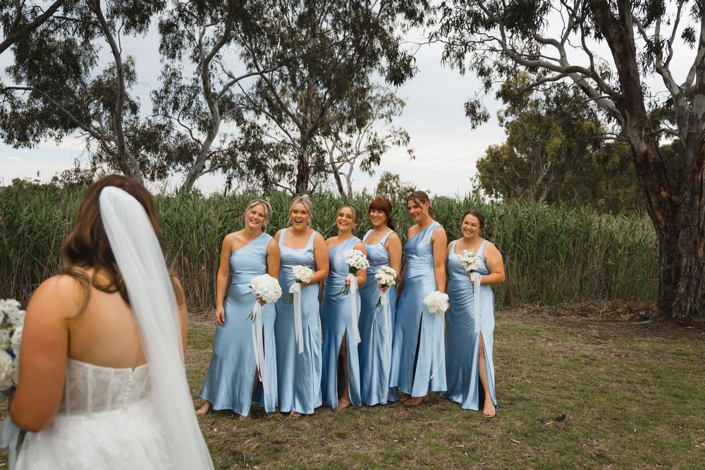A bride in white stands with a group wearing matching light blue bridesmaid dresses in a natural outdoor setting.
