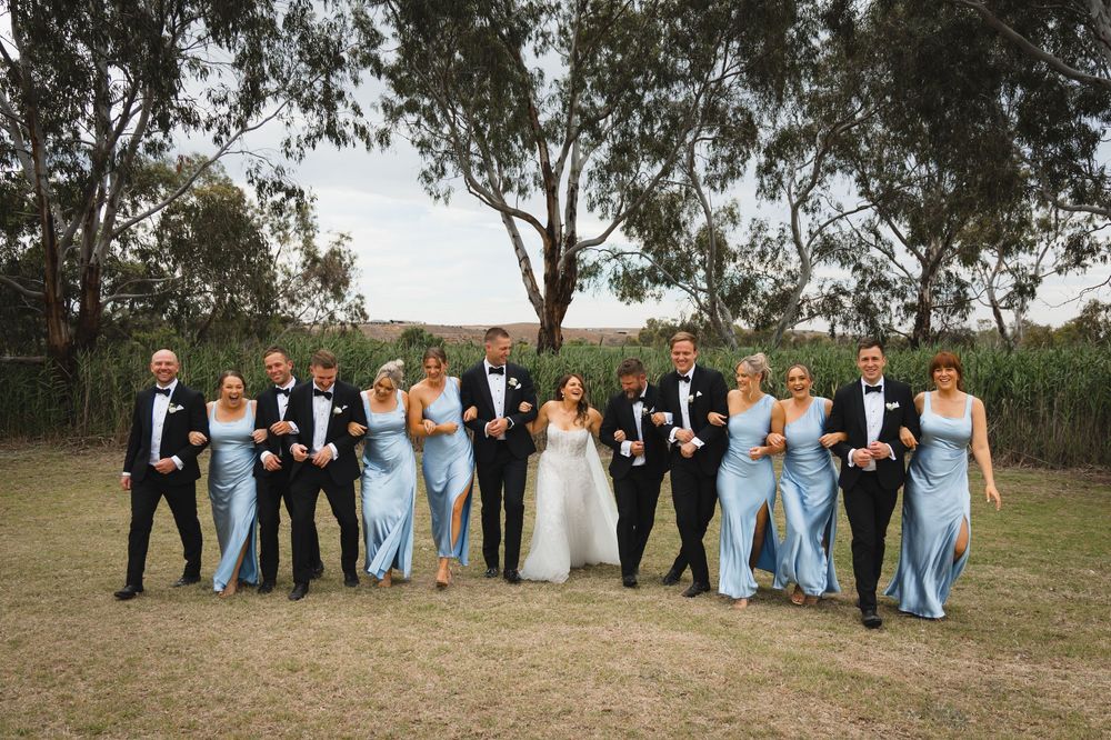 Wedding party wearing black tuxedos and light blue bridesmaid dresses pose together outdoors under gum trees.