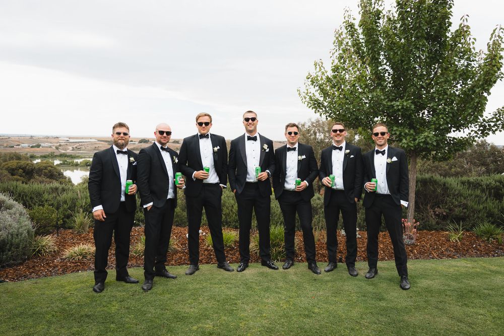 A group of groomsmen in black tuxedos stand together on grass with a scenic backdrop.