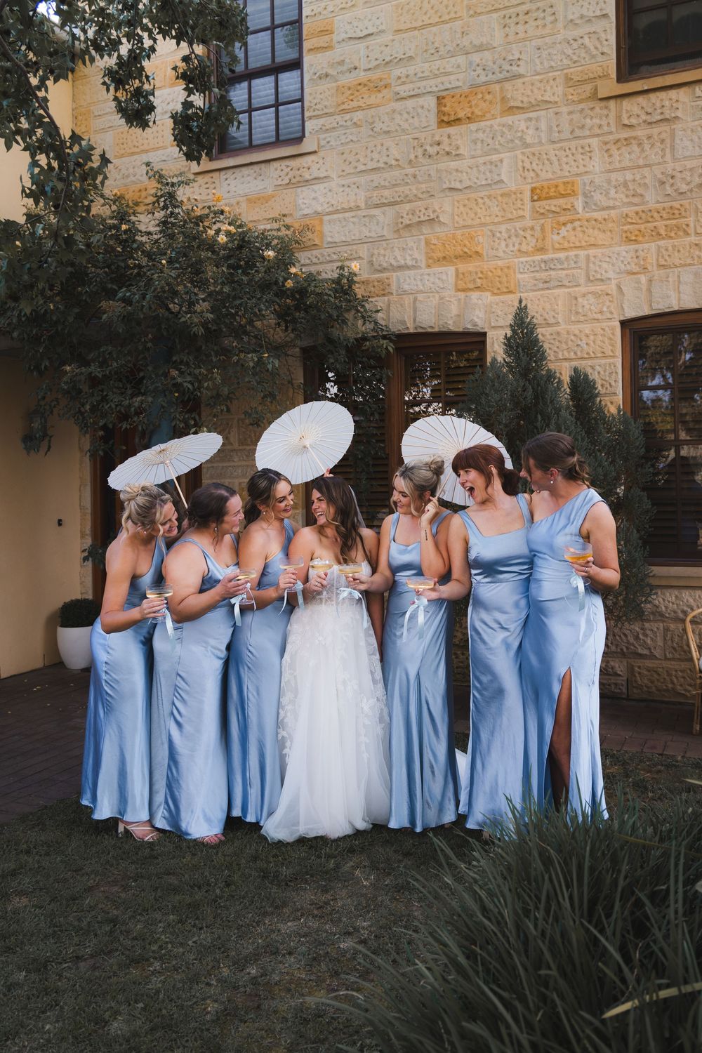 A bridal party in light blue dresses holding white parasols gather outside a stone building during a wedding celebration.