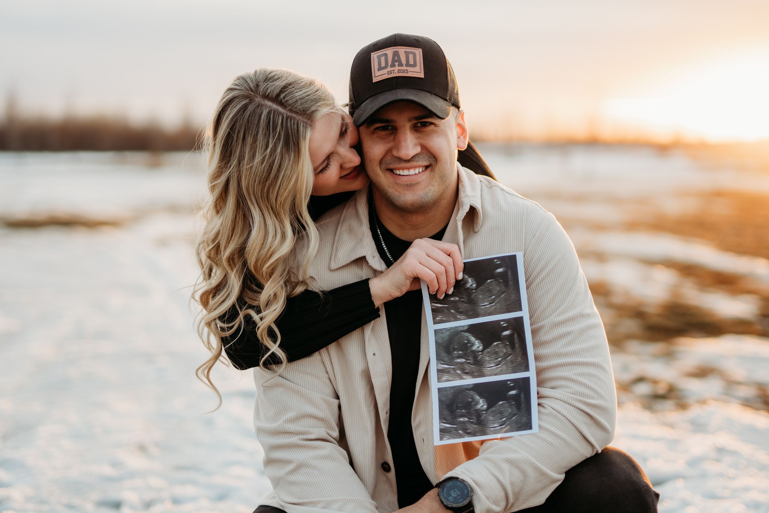 A couple shares an intimate moment on a beach at sunset while holding an ultrasound photo. Fort McMurray
