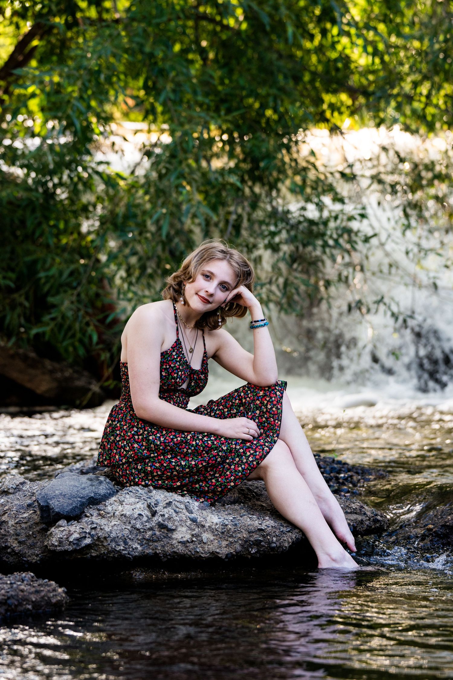 Person in floral dress sits on rock by rushing waterfall under green trees in dreamy nature portrait setting.