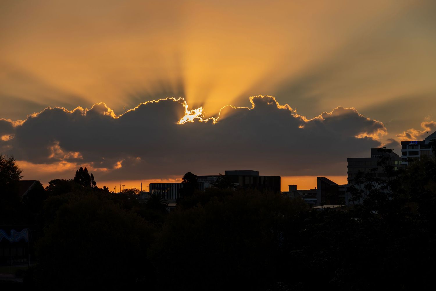 Golden sunset casting radiant beams from behind dramatic clouds over silhouetted rooftops and treetops.
