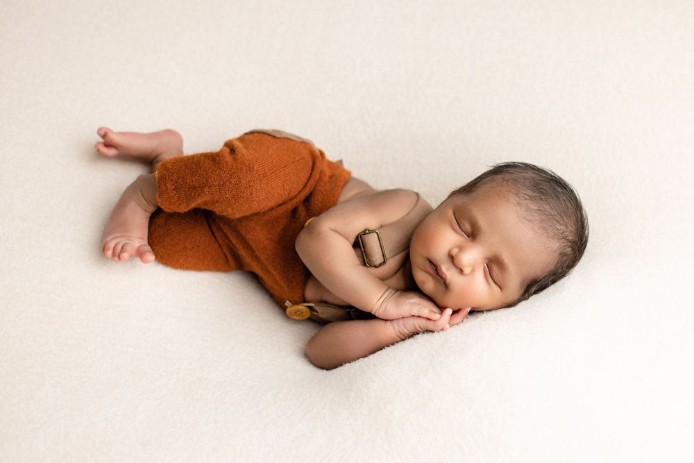 A sleeping newborn baby wearing rust-colored pants rests peacefully on a cream-colored backdrop.