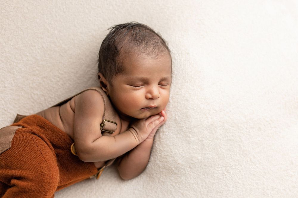 A sleeping baby wearing orange pants lies on a light-colored blanket with one hand under their chin.