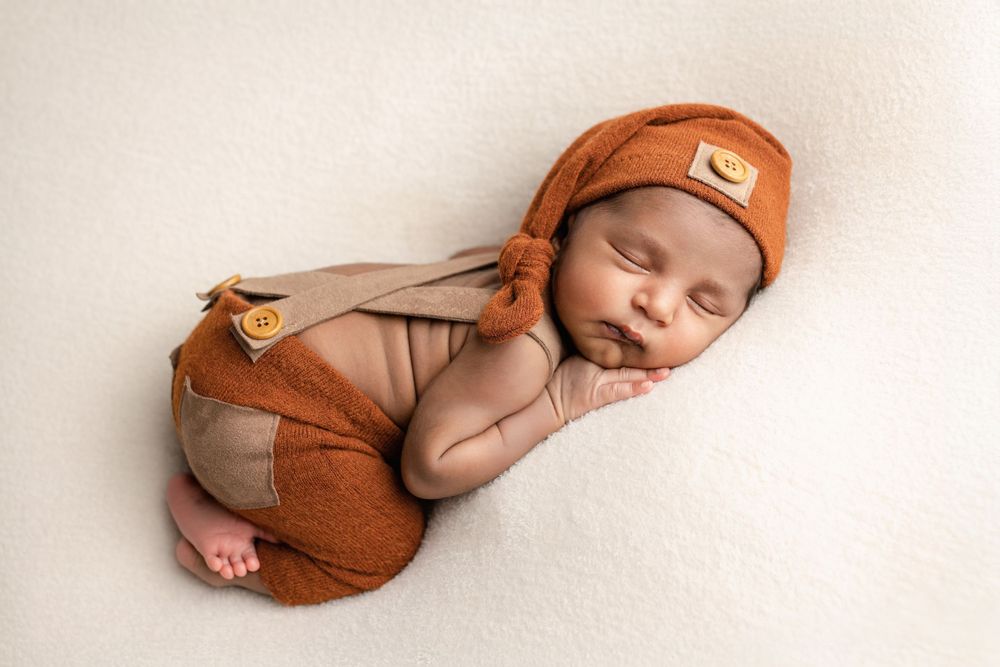 A sleeping newborn in a brown knit cap rests peacefully on white fabric while wearing cozy brown clothing.
